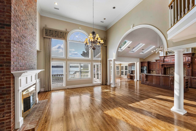 unfurnished living room with hardwood / wood-style flooring, high vaulted ceiling, a brick fireplace, crown molding, and ornate columns