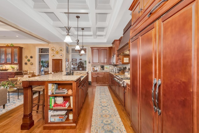 kitchen with a breakfast bar, decorative light fixtures, an island with sink, coffered ceiling, and light stone countertops