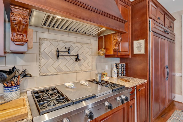 kitchen with light stone counters, paneled built in fridge, custom range hood, stainless steel gas cooktop, and decorative backsplash