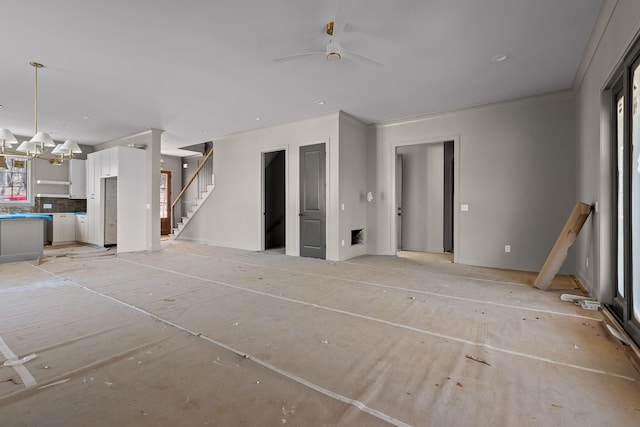 unfurnished living room featuring ornamental molding, ceiling fan with notable chandelier, and stairs