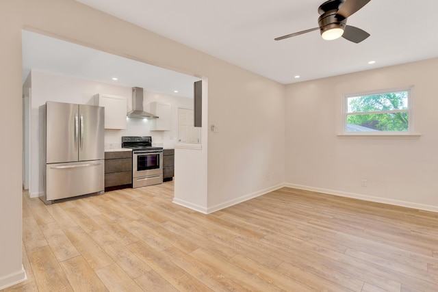 kitchen featuring wall chimney range hood, appliances with stainless steel finishes, ceiling fan, and light wood-style flooring