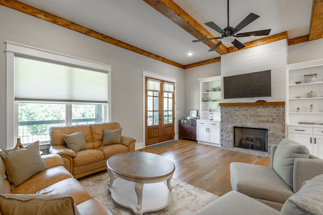 living room with beam ceiling, ceiling fan, light wood-type flooring, a brick fireplace, and built in shelves