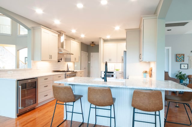 kitchen with wall chimney exhaust hood, wine cooler, a breakfast bar, white cabinetry, and light stone counters