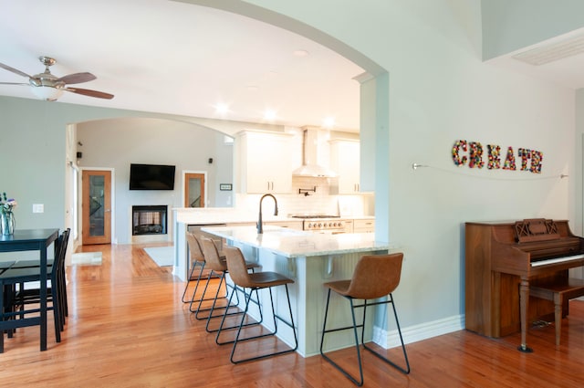 kitchen with wall chimney exhaust hood, light hardwood / wood-style flooring, kitchen peninsula, white cabinets, and light stone counters