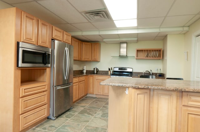 kitchen with appliances with stainless steel finishes, wall chimney exhaust hood, a paneled ceiling, and light brown cabinets