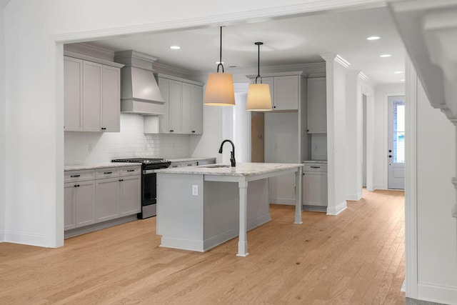 kitchen with light wood-type flooring, custom exhaust hood, stainless steel gas range, pendant lighting, and an island with sink