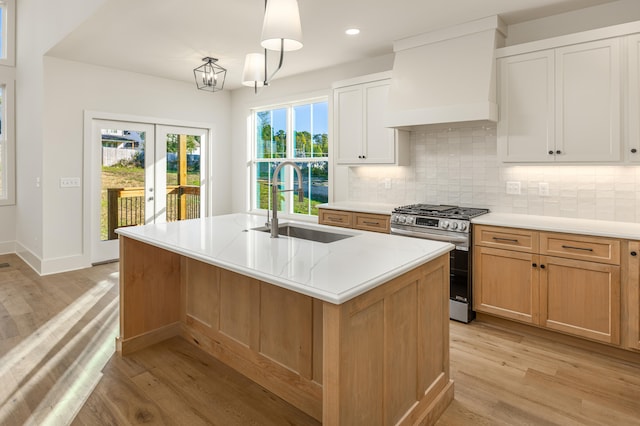 kitchen with light wood-type flooring, stainless steel range with gas cooktop, white cabinets, premium range hood, and a center island with sink