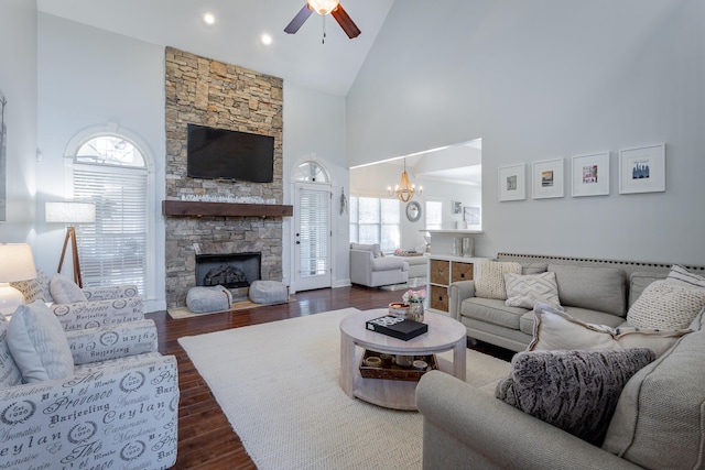 living room with high vaulted ceiling, dark wood-type flooring, a stone fireplace, and ceiling fan with notable chandelier