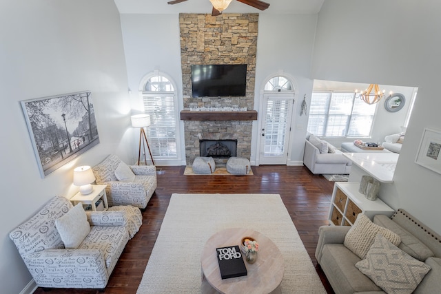 living room featuring dark wood-type flooring, a towering ceiling, a stone fireplace, and ceiling fan with notable chandelier