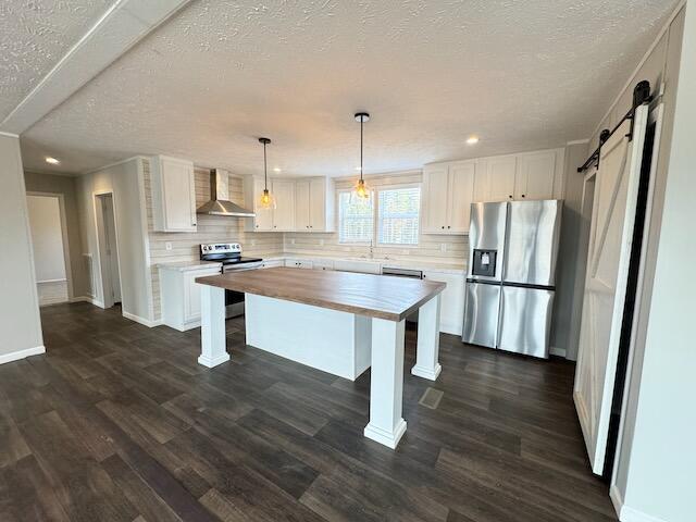 kitchen featuring a barn door, wood counters, stainless steel appliances, wall chimney range hood, and white cabinets