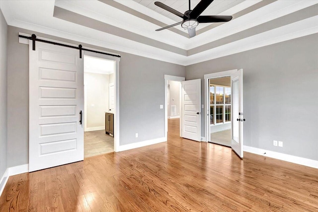 unfurnished bedroom with ceiling fan, a raised ceiling, a barn door, connected bathroom, and light hardwood / wood-style flooring