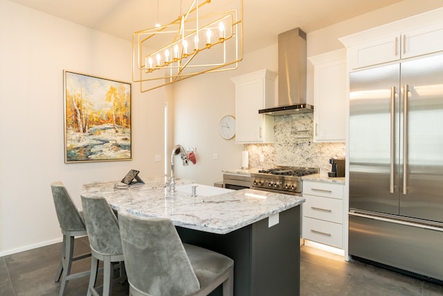 kitchen with appliances with stainless steel finishes, an island with sink, hanging light fixtures, wall chimney exhaust hood, and white cabinets