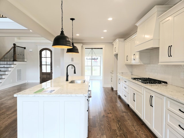 kitchen featuring sink, premium range hood, light stone counters, an island with sink, and decorative light fixtures