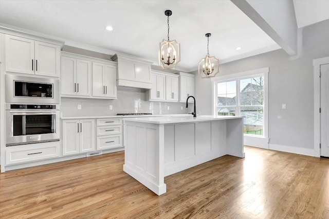 kitchen with a kitchen island with sink, stainless steel oven, white cabinets, light countertops, and decorative light fixtures