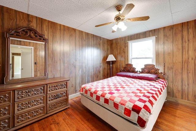 bedroom featuring wooden walls, a drop ceiling, wood-type flooring, and ceiling fan