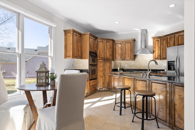 kitchen featuring wall chimney exhaust hood, crown molding, stainless steel appliances, and dark stone counters