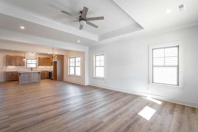 unfurnished living room with crown molding, a raised ceiling, ceiling fan with notable chandelier, and light wood-type flooring