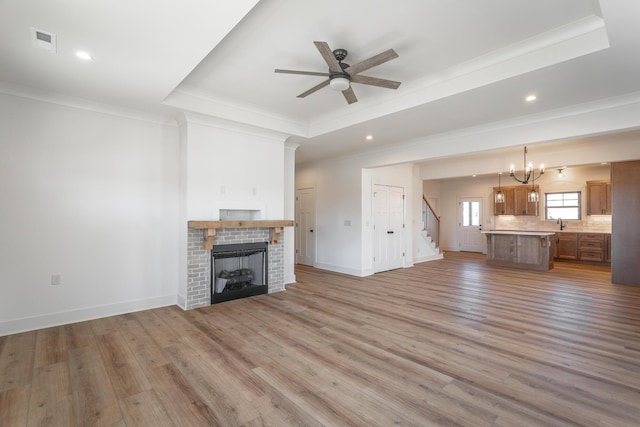 unfurnished living room featuring crown molding, a tray ceiling, a brick fireplace, ceiling fan with notable chandelier, and light wood-type flooring