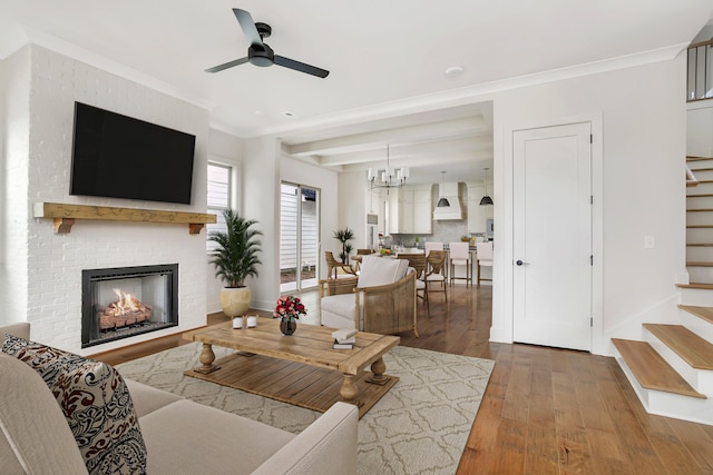 living room featuring a brick fireplace, ceiling fan with notable chandelier, dark wood-type flooring, crown molding, and beamed ceiling