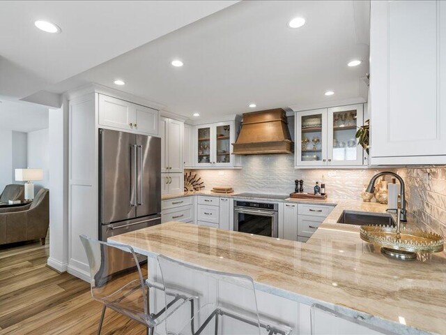 kitchen with white cabinetry, light hardwood / wood-style floors, kitchen peninsula, custom exhaust hood, and stainless steel appliances