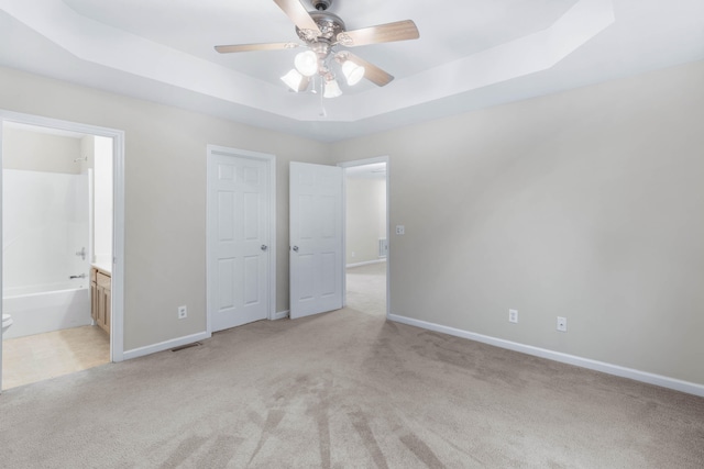 unfurnished bedroom with a tray ceiling, ensuite bath, ceiling fan, and light colored carpet