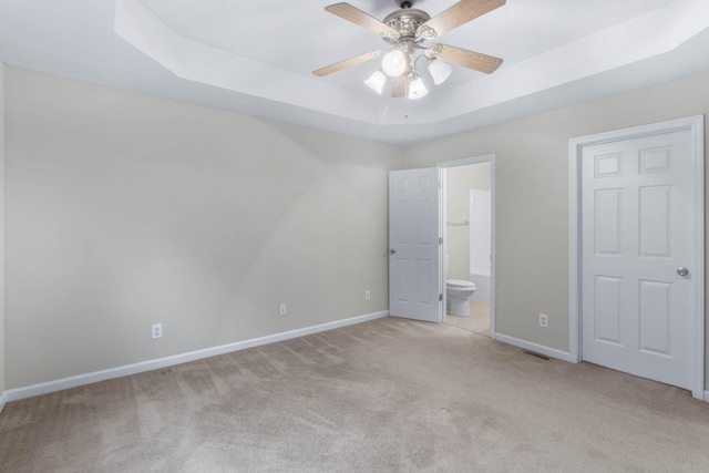 unfurnished bedroom with light carpet, a tray ceiling, ceiling fan, and ensuite bathroom