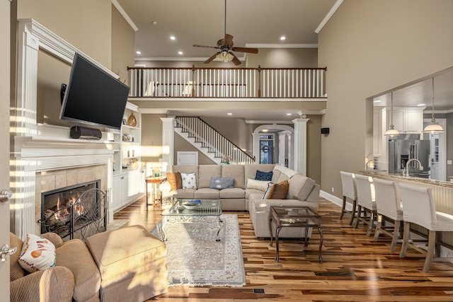 living room with a high ceiling, ceiling fan, ornamental molding, wood-type flooring, and a tiled fireplace