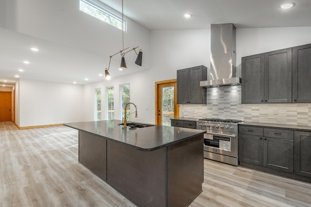 kitchen with sink, wall chimney exhaust hood, light wood-type flooring, high end stove, and an island with sink