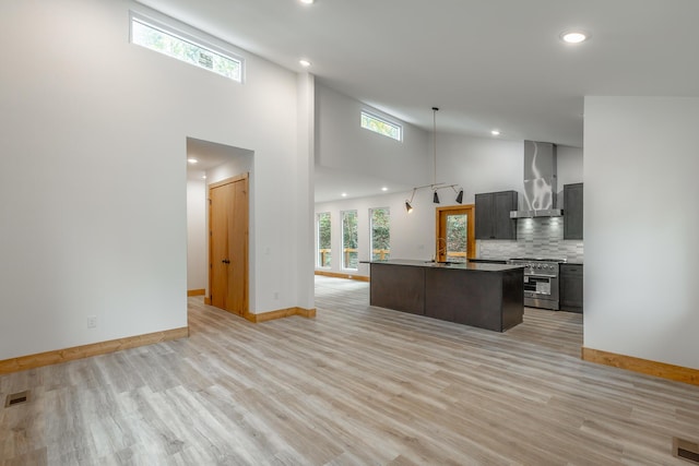 kitchen with a high ceiling, a center island with sink, stainless steel stove, wall chimney exhaust hood, and light wood-type flooring