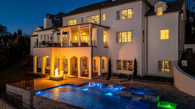 back house at night featuring a balcony, a fire pit, a patio area, and a pool with hot tub