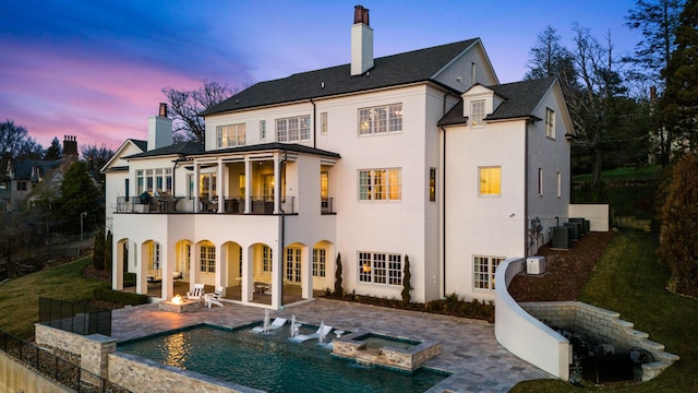 back house at dusk featuring a fire pit, cooling unit, a pool with hot tub, a balcony, and a patio