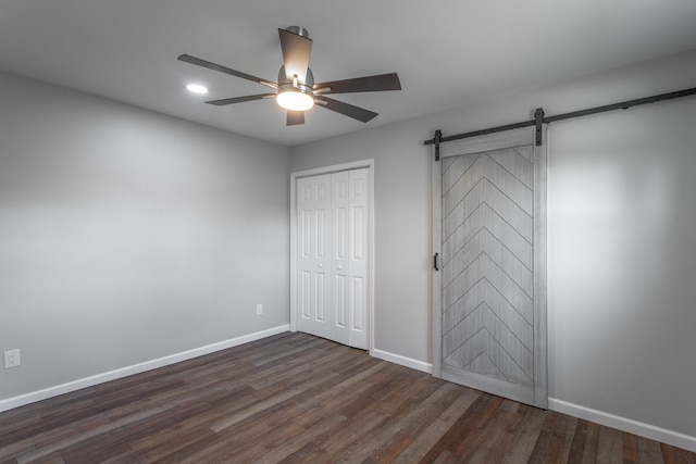 unfurnished bedroom featuring ceiling fan, a barn door, dark wood-type flooring, and a closet