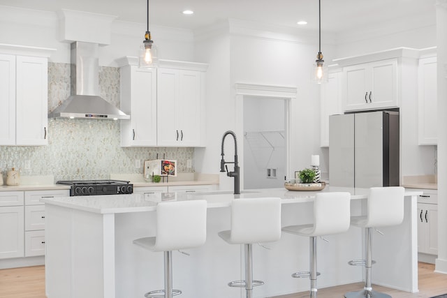 kitchen with a center island with sink, white cabinetry, hanging light fixtures, and wall chimney range hood