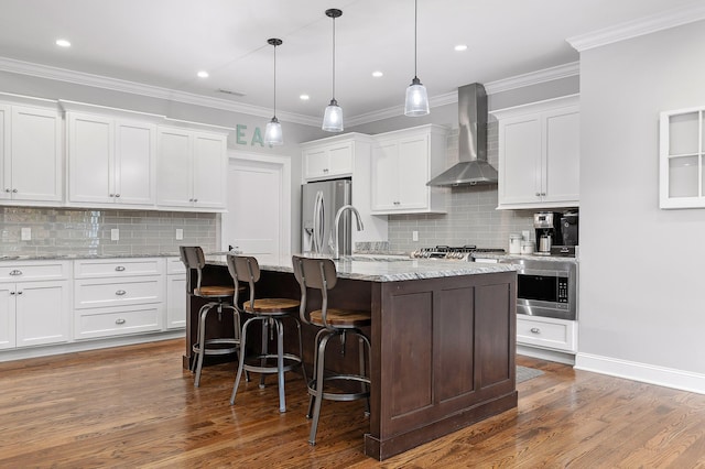 kitchen with appliances with stainless steel finishes, wall chimney exhaust hood, white cabinets, and an island with sink