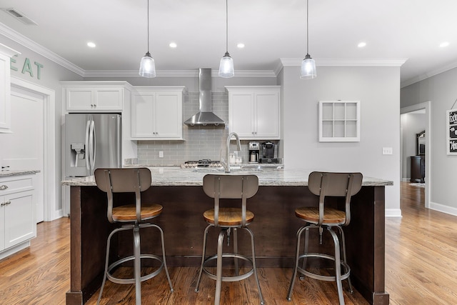 kitchen with a large island with sink, wall chimney exhaust hood, decorative light fixtures, white cabinetry, and stainless steel fridge with ice dispenser