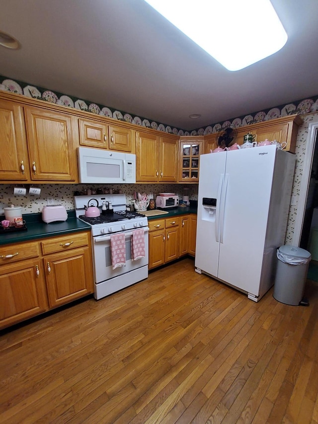 kitchen featuring dark countertops, glass insert cabinets, brown cabinetry, light wood-type flooring, and white appliances