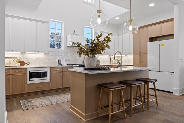 kitchen with stainless steel microwave, dark wood-type flooring, decorative backsplash, pendant lighting, and a kitchen island with sink