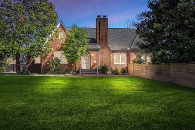 back of property at dusk with a lawn, brick siding, a fenced backyard, and roof with shingles