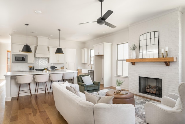 living room with ceiling fan, sink, a brick fireplace, hardwood / wood-style floors, and ornamental molding