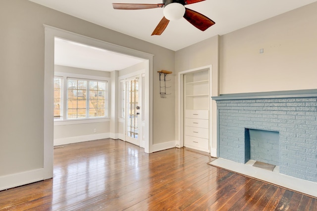 unfurnished living room with built in shelves, a ceiling fan, wood-type flooring, baseboards, and a brick fireplace