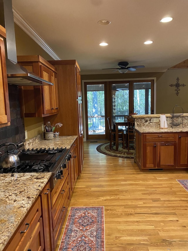 kitchen featuring stainless steel gas stovetop, wall chimney exhaust hood, ceiling fan, light stone countertops, and ornamental molding