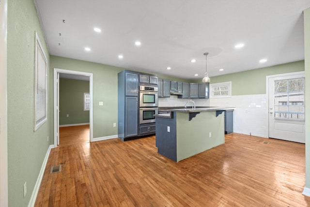 kitchen with double oven, wood-type flooring, decorative light fixtures, a breakfast bar area, and a kitchen island with sink