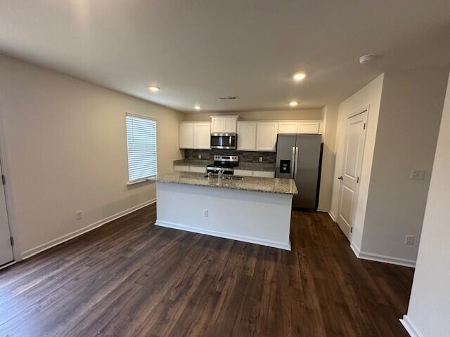 kitchen with appliances with stainless steel finishes, dark hardwood / wood-style flooring, light stone counters, white cabinets, and an island with sink