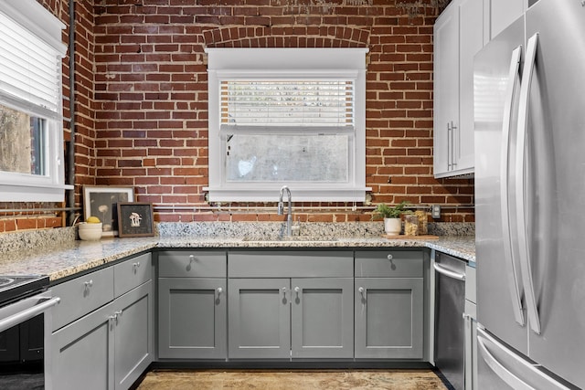 kitchen featuring gray cabinetry, sink, light stone countertops, stainless steel appliances, and brick wall