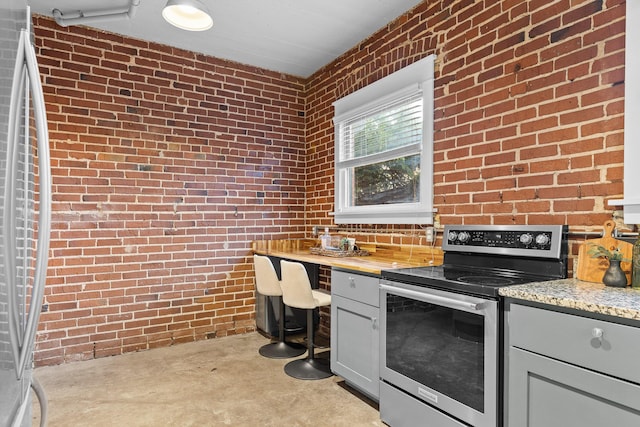 kitchen featuring gray cabinetry, stainless steel electric range oven, light stone counters, brick wall, and fridge