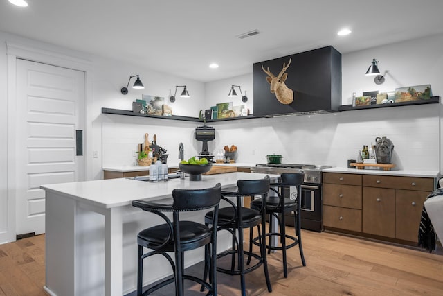 kitchen featuring stainless steel stove, wall chimney exhaust hood, decorative backsplash, light wood-type flooring, and dark brown cabinetry