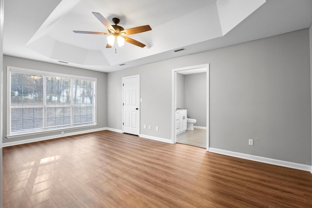 unfurnished bedroom with ceiling fan, a raised ceiling, wood-type flooring, and connected bathroom