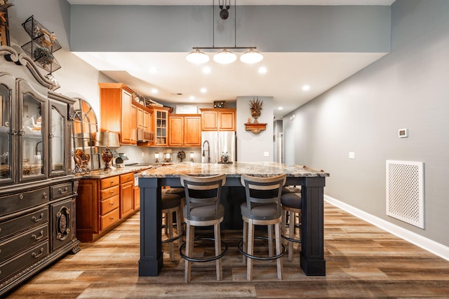 kitchen with light stone countertops, stainless steel fridge, a kitchen island with sink, pendant lighting, and light hardwood / wood-style floors