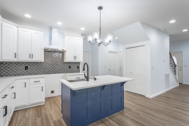 kitchen featuring hanging light fixtures, white cabinets, wall chimney range hood, a center island with sink, and an inviting chandelier