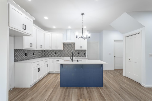 kitchen featuring hanging light fixtures, wall chimney exhaust hood, white cabinetry, a kitchen island with sink, and light hardwood / wood-style floors